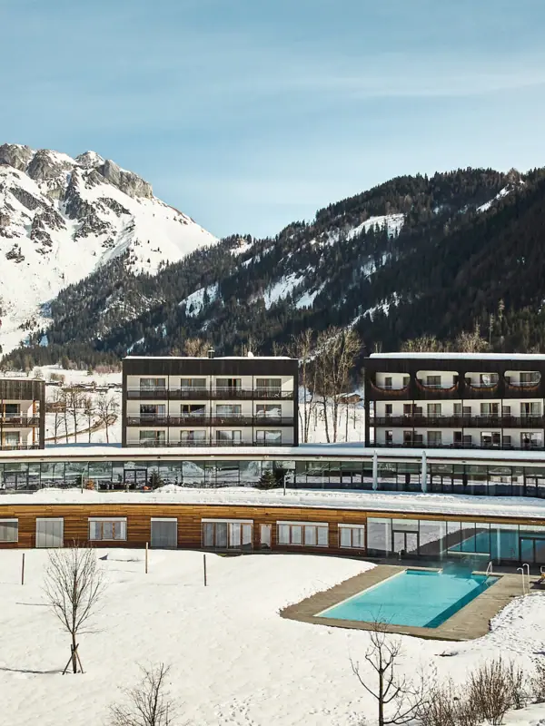 The hotel building with its outdoor swimming pool in the snow, surrounded by mountains.