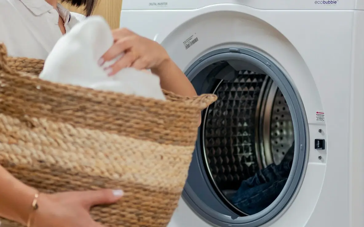 Woman holding a laundry basket and standing next to a washing machine.