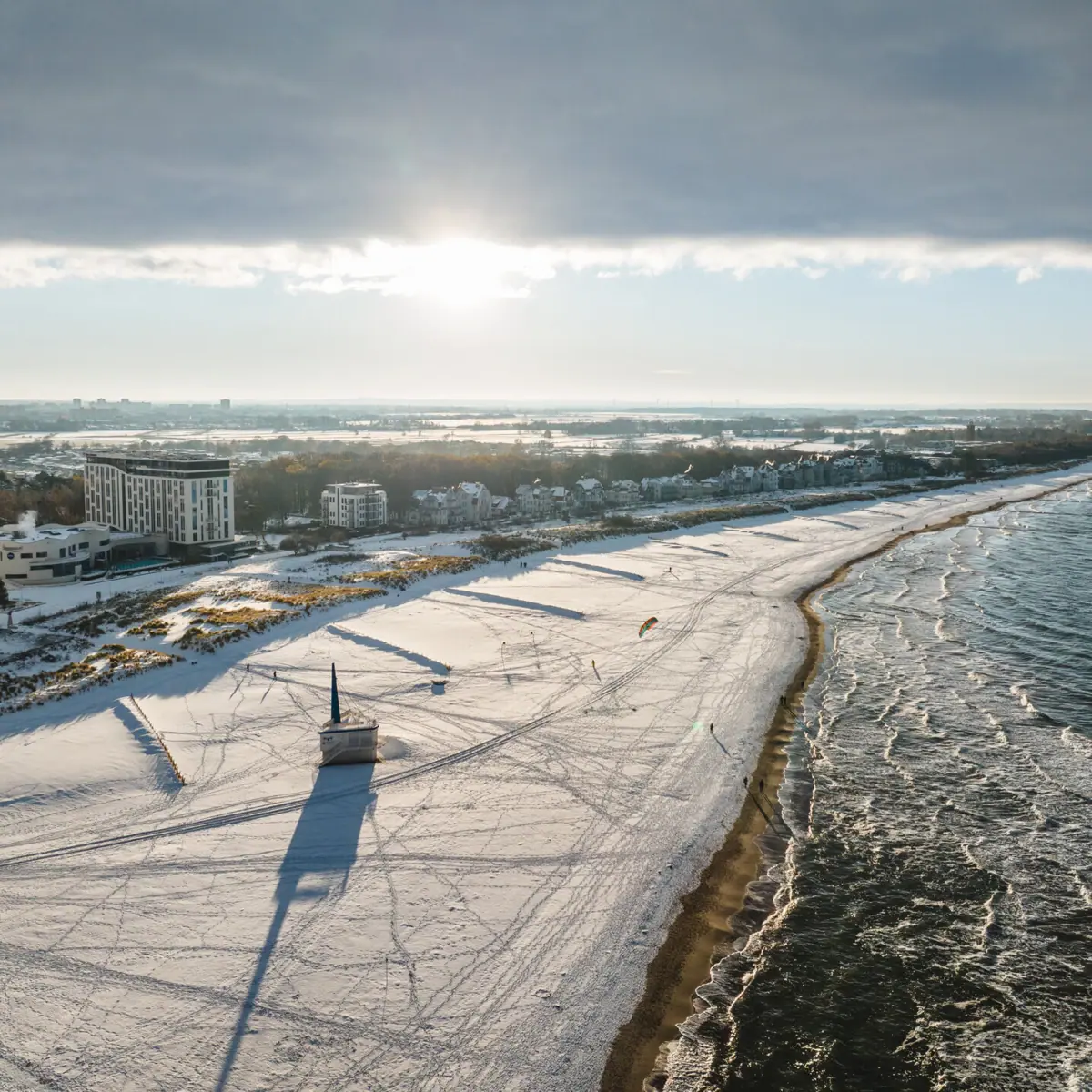A snow-covered beach in Warnemünde in winter, with buildings and water in the background.