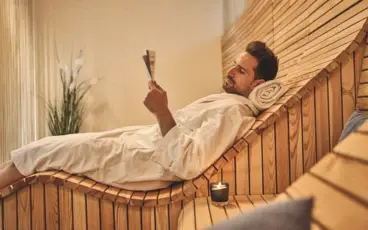 A man in a bathrobe lies on a wooden lounger and reads a desk.