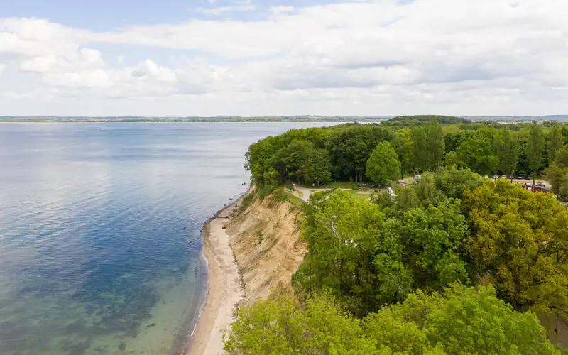 Scenic coastal view featuring a steep cliff lined with lush green trees, a narrow sandy beach, and calm blue waters under a partly cloudy sky.