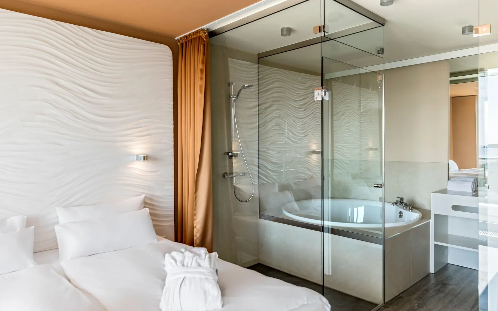 Modern hotel room with a comfortable double bed, stylish wave-patterned wall design, and an open-plan bathroom with glass partitions and a bathtub, creating a bright and luxurious atmosphere.