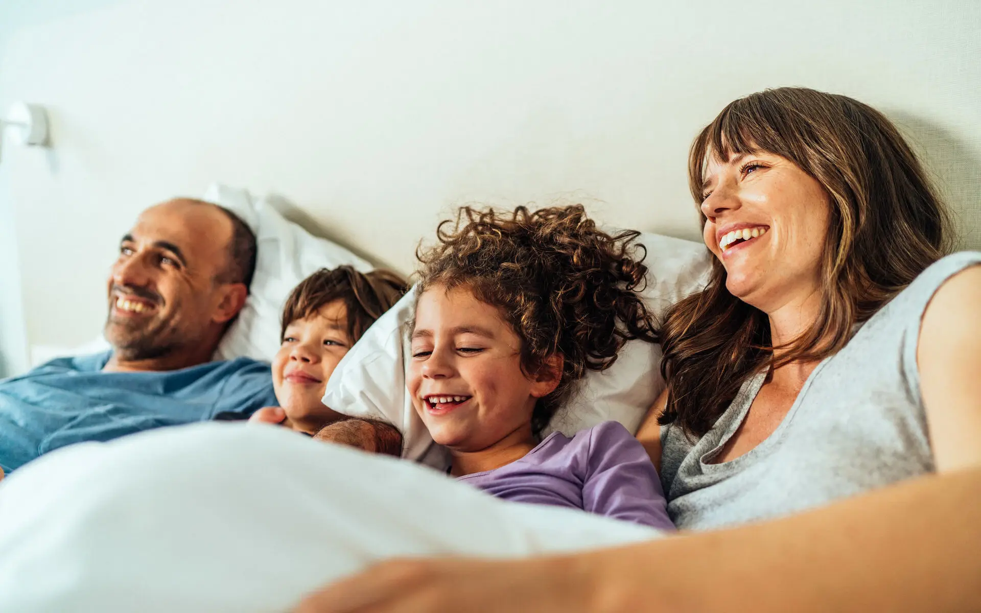 A family of four lie together in a bed and laugh