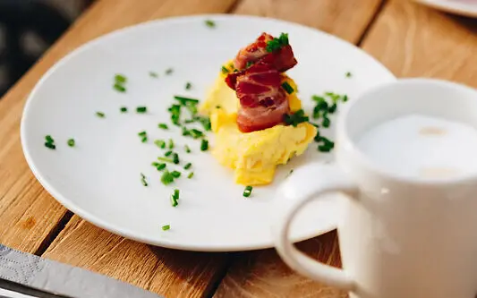 A plate of scrambled eggs and bacon on a table, surrounded by cutlery and a cup of coffee.