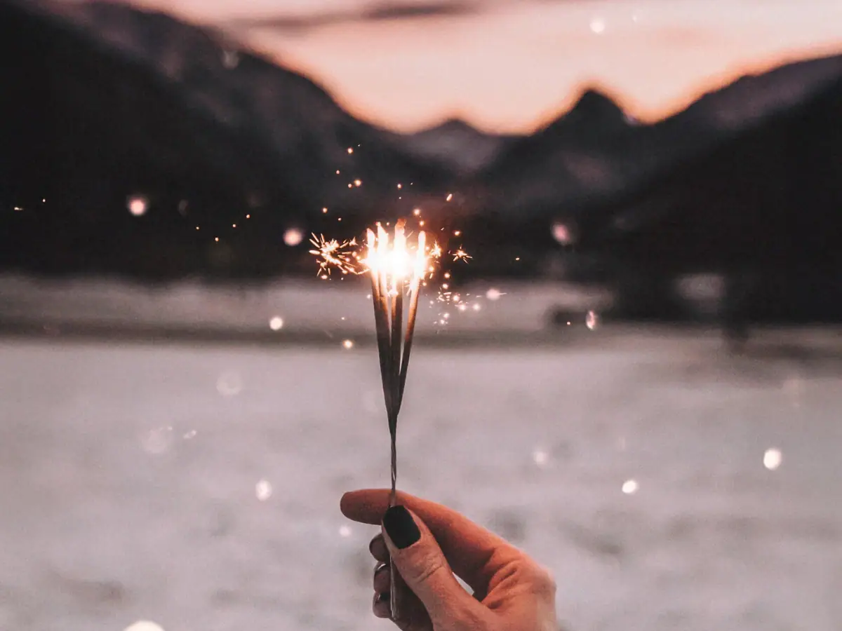 A hand is holding a sparkler against a sky with mountains in the background