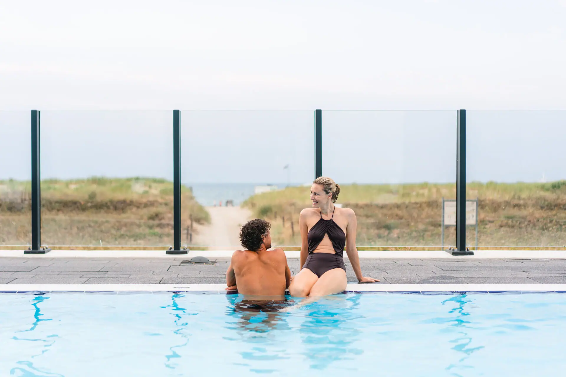 A man and a woman sit in a swimming pool, with dunes and the sea in the background.