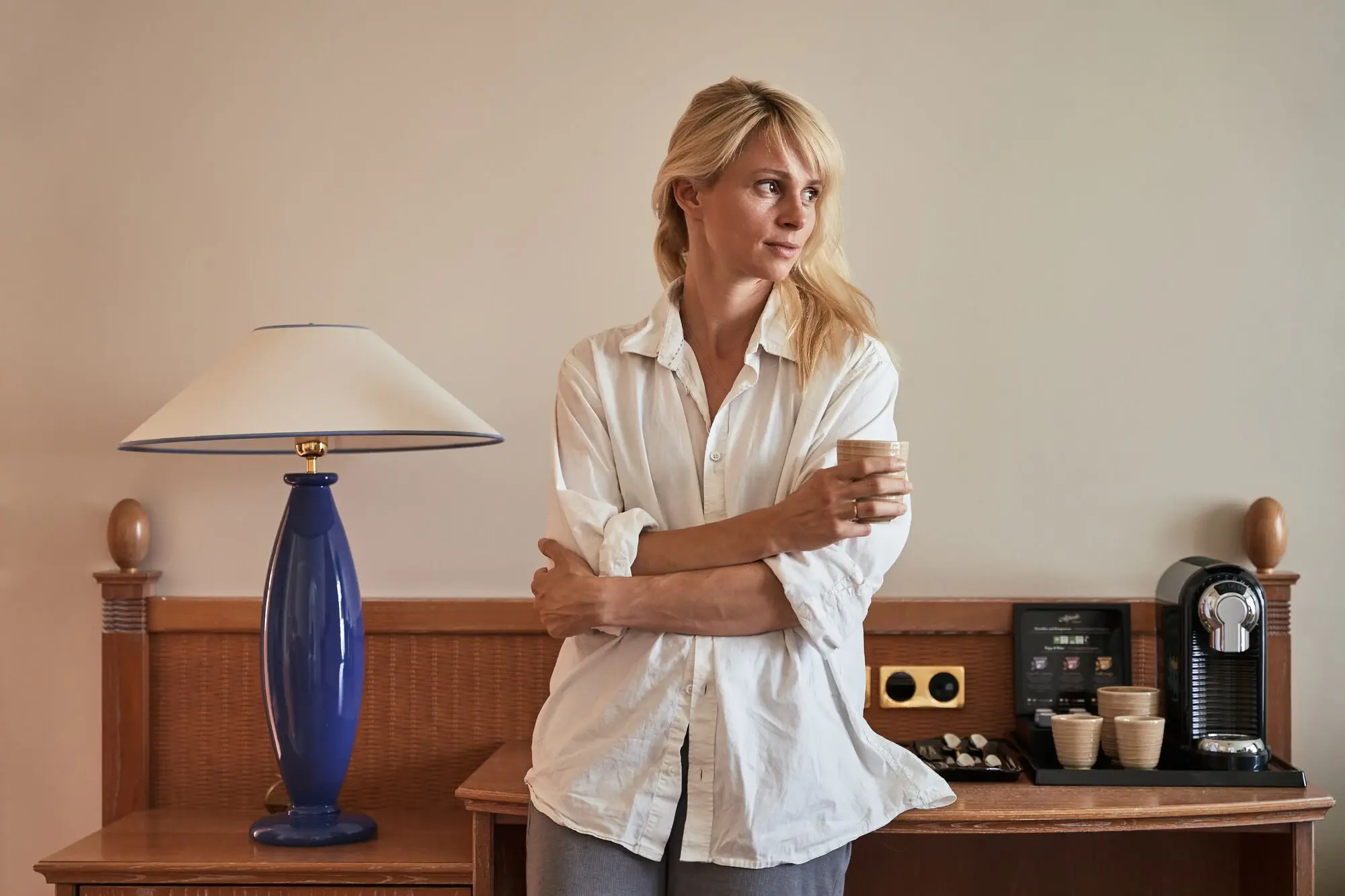 Woman standing in front of a table with a lamp and a coffee machine, holding a mug in her hand.