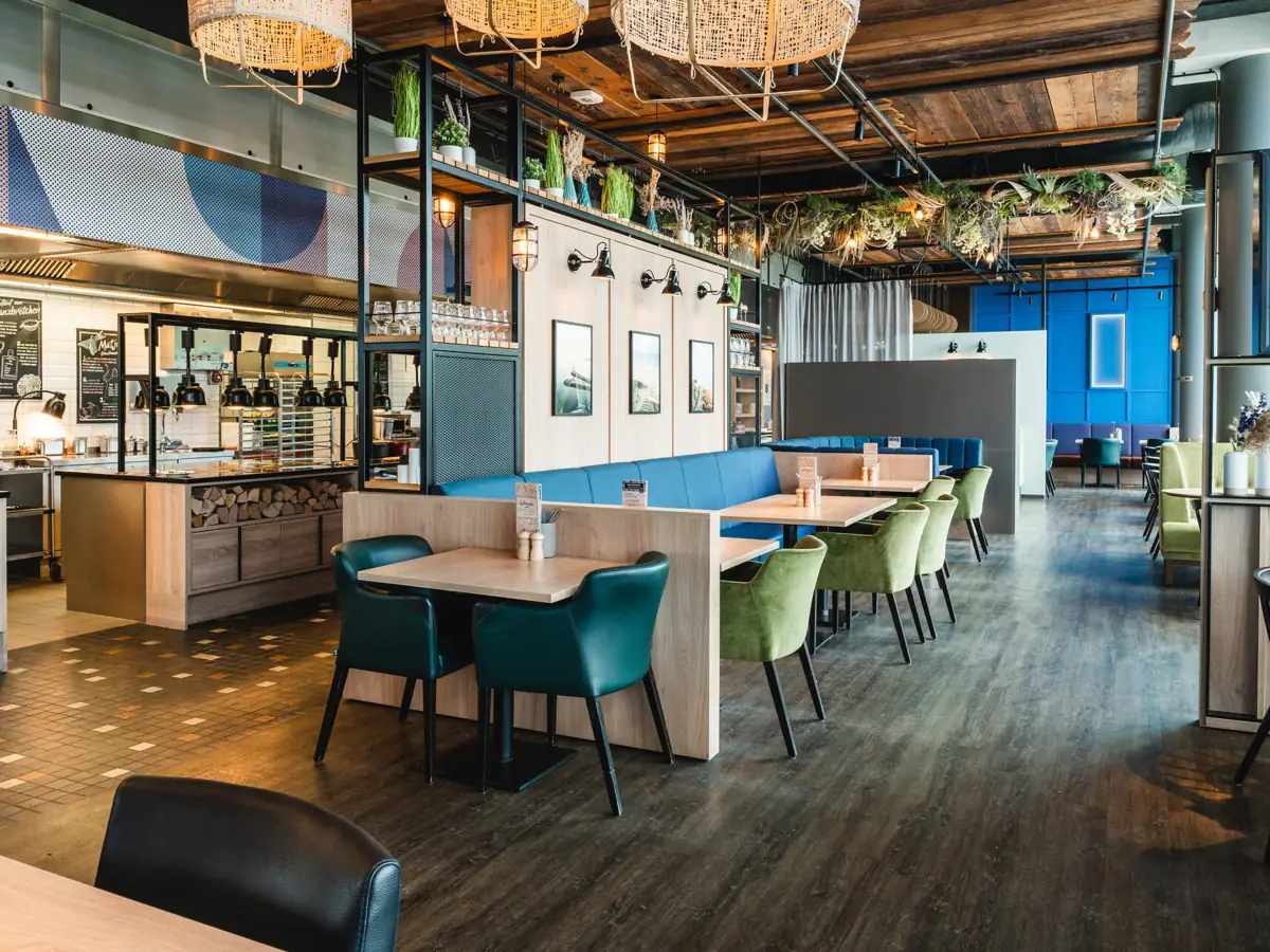 A restaurant with tables and chairs. Stylish and modern furnishings. Maritime look with pictures of Warnemünde on the wall. Open buffet area in the background.