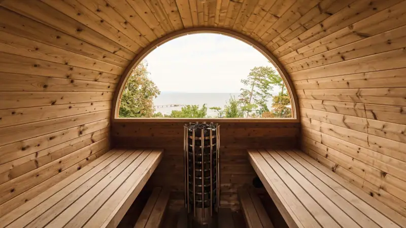 A barrel sauna from the inside with a round window overlooking the water.