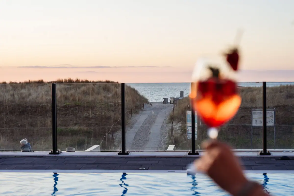 A hand is holding a wine glass filled with strawberries above a swimming pool.