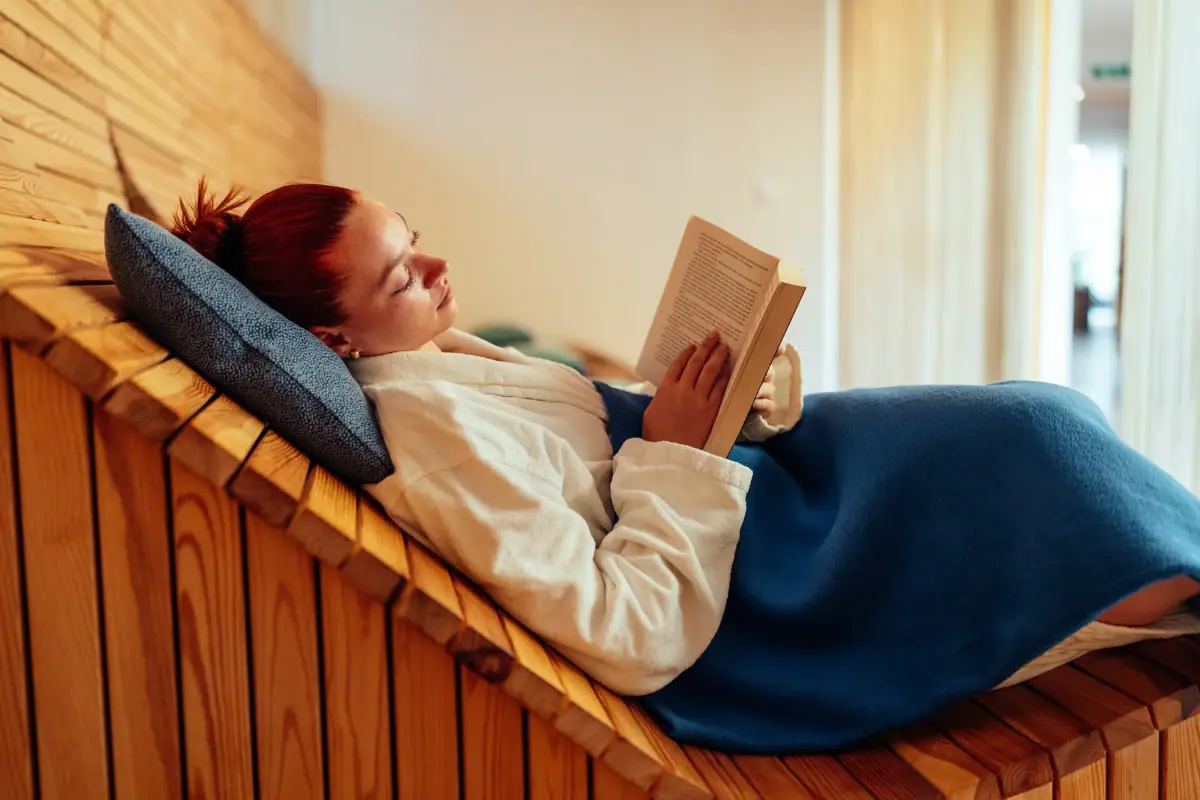 A woman is lying on a wooden bench, reading a book.