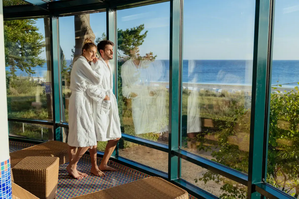 A man and a woman in bathrobes are standing in a room with large windows.